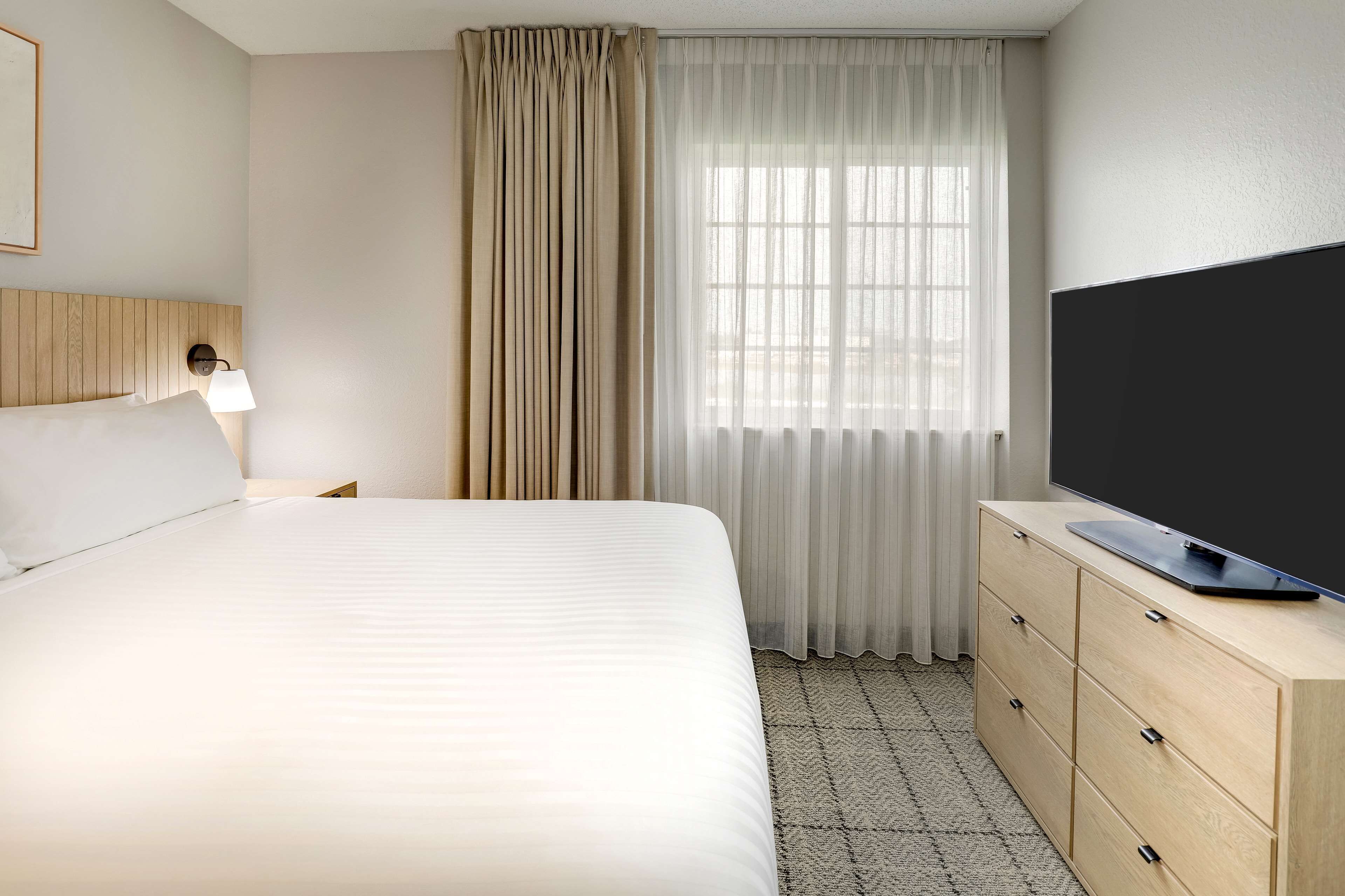 Habitación de hotel con una cama grande, un televisor sobre una cómoda de madera, una lámpara y un ventanal con cortinas translúcidas.