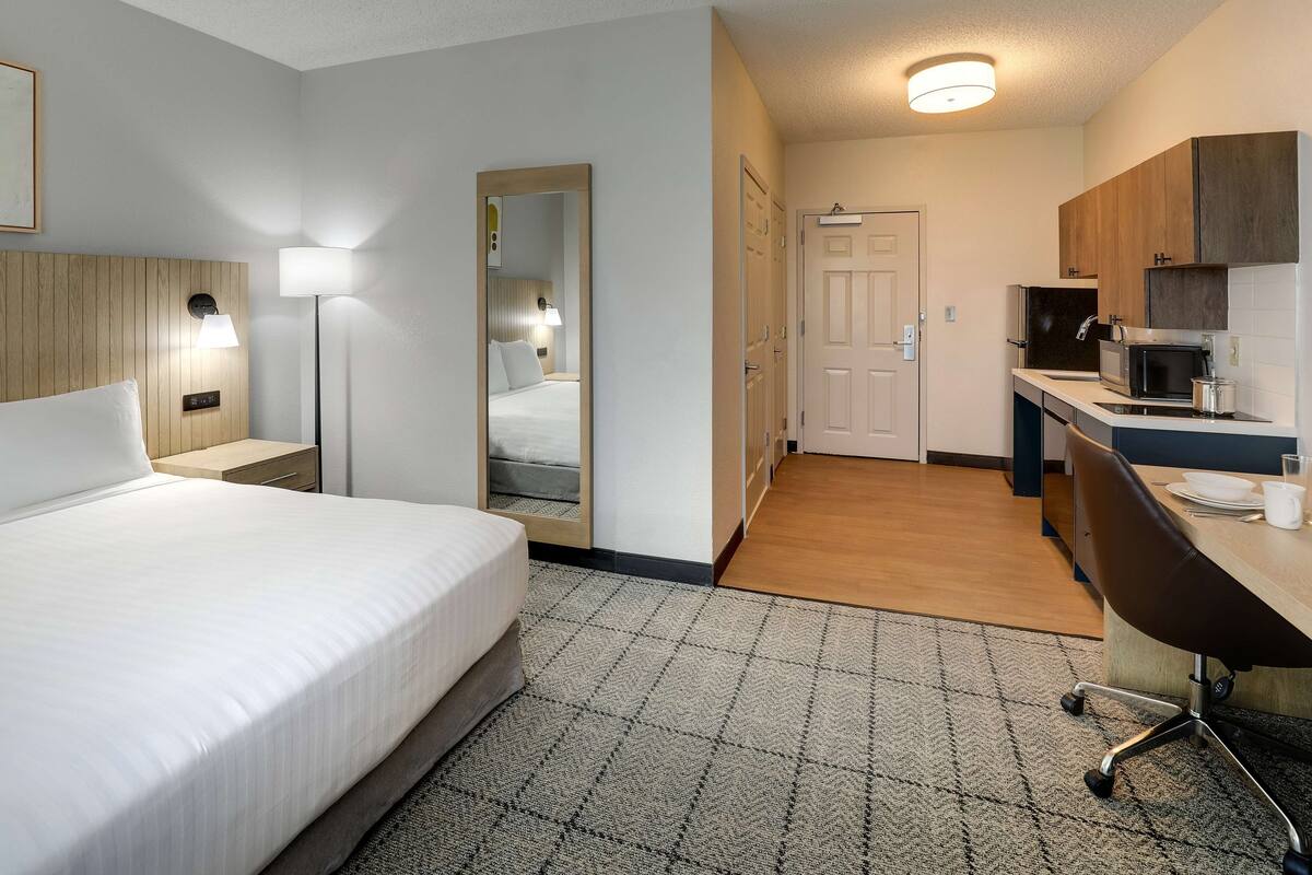 A hotel room with a bed, bedside table, lamp, mirror, and a kitchenette with a microwave, toaster, and coffee maker.