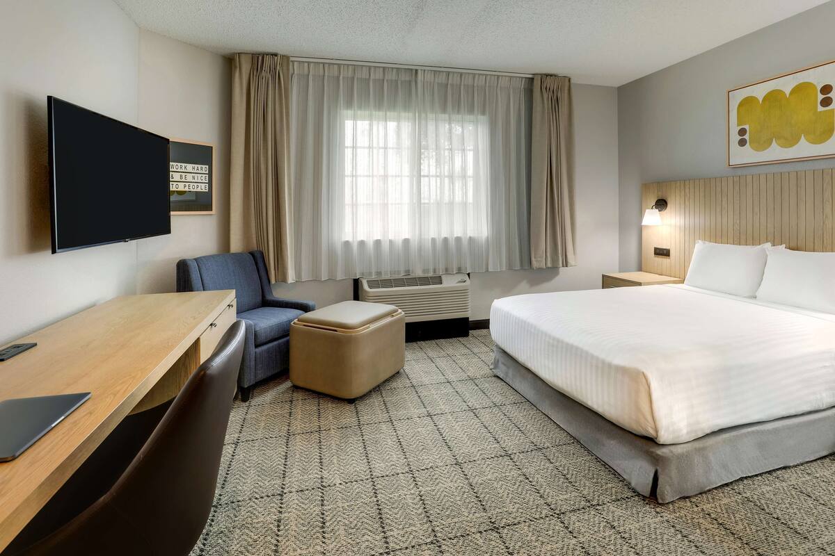 A hotel room with a bed, a desk, a chair, a TV, a window with curtains, and a wall with a motivational quote.