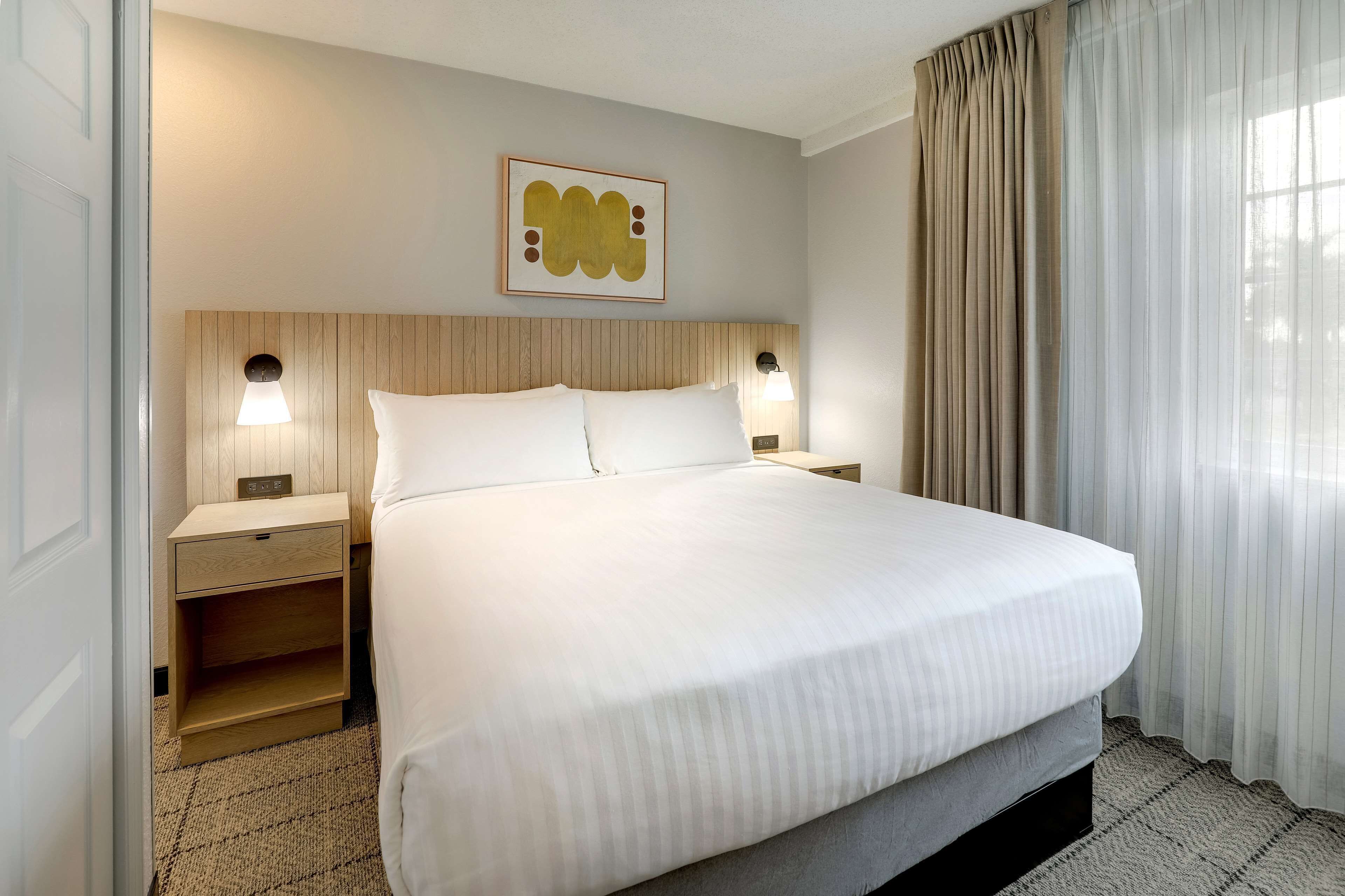 Una habitación de hotel con una cama grande, dos mesitas de noche con lámparas, un cabecero de madera y una obra de arte enmarcada en la pared.
