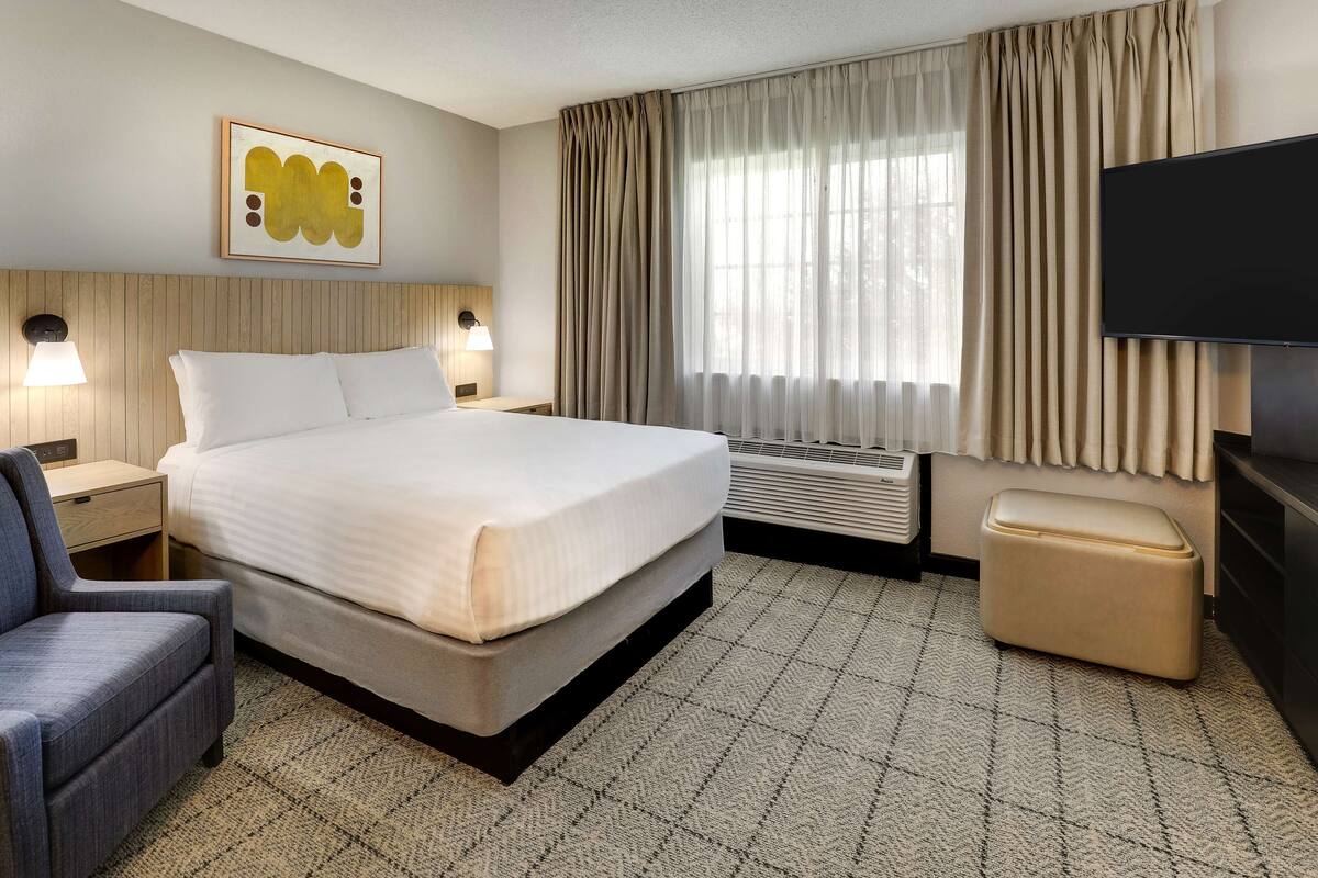 A hotel room with a large bed, a desk, a chair, a TV, and a window with curtains.