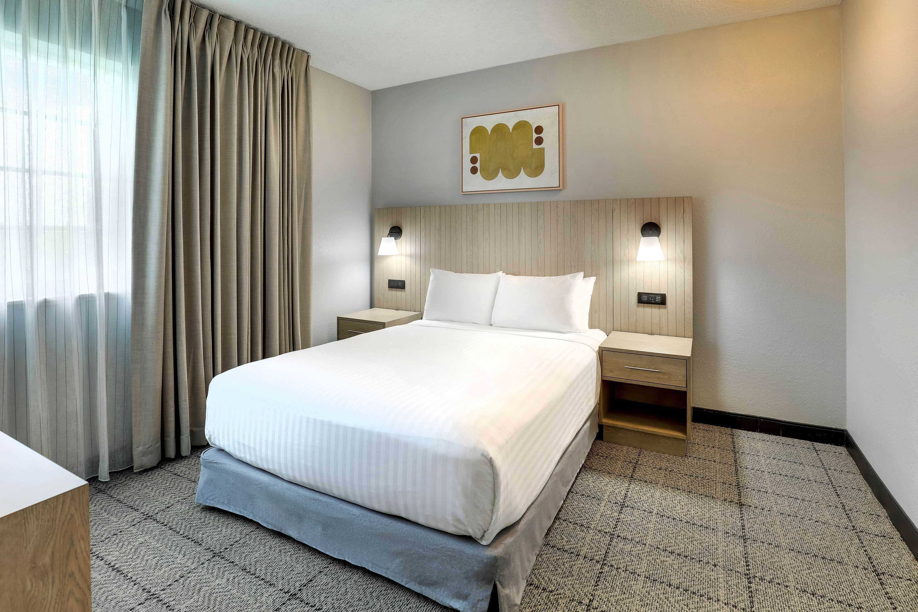 A hotel room with a large bed, two bedside tables, a headboard with a light fixture, and a framed picture on the wall.