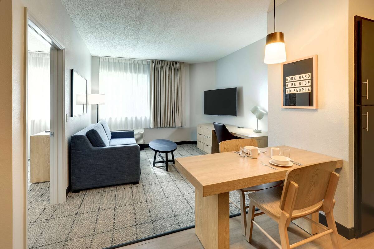 A modern hotel room with a sofa, a dining table, a TV, and a motivational poster.