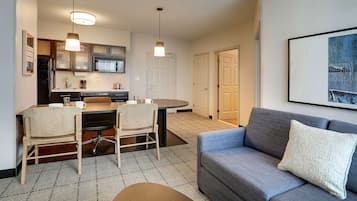 A modern living room with a gray sofa, a dining table with chairs, and a kitchen area with a refrigerator and microwave.
