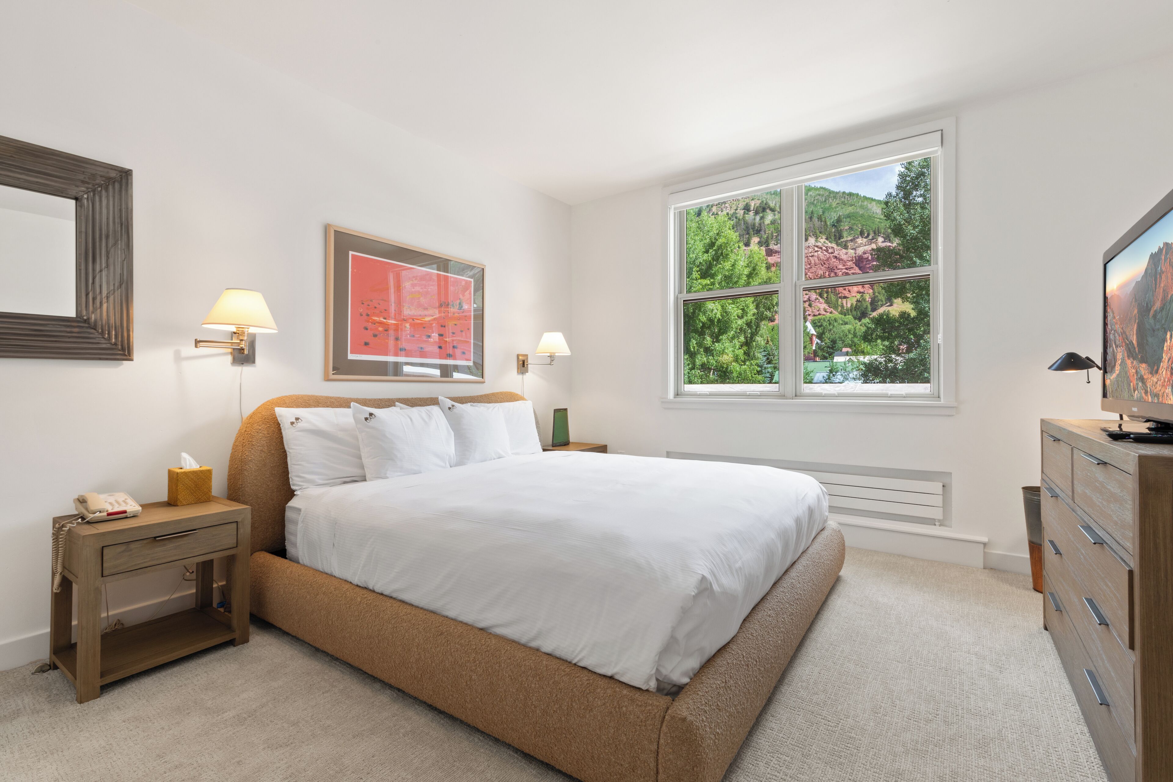 A bedroom with a large bed, bedside lamps, a nightstand, a dresser, and a window with a view of greenery.