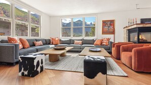 A modern living room with a grey sofa set, orange cushions, a fireplace, and a large window with a mountain view. - Camel's Garden Hotel (Telluride)