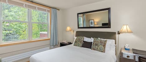A hotel room with a large bed, a desk with a chair, a nightstand with a lamp, and a window with curtains.