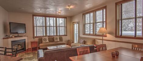 A spacious living room with a fireplace, large windows, a dining table, and comfortable seating.