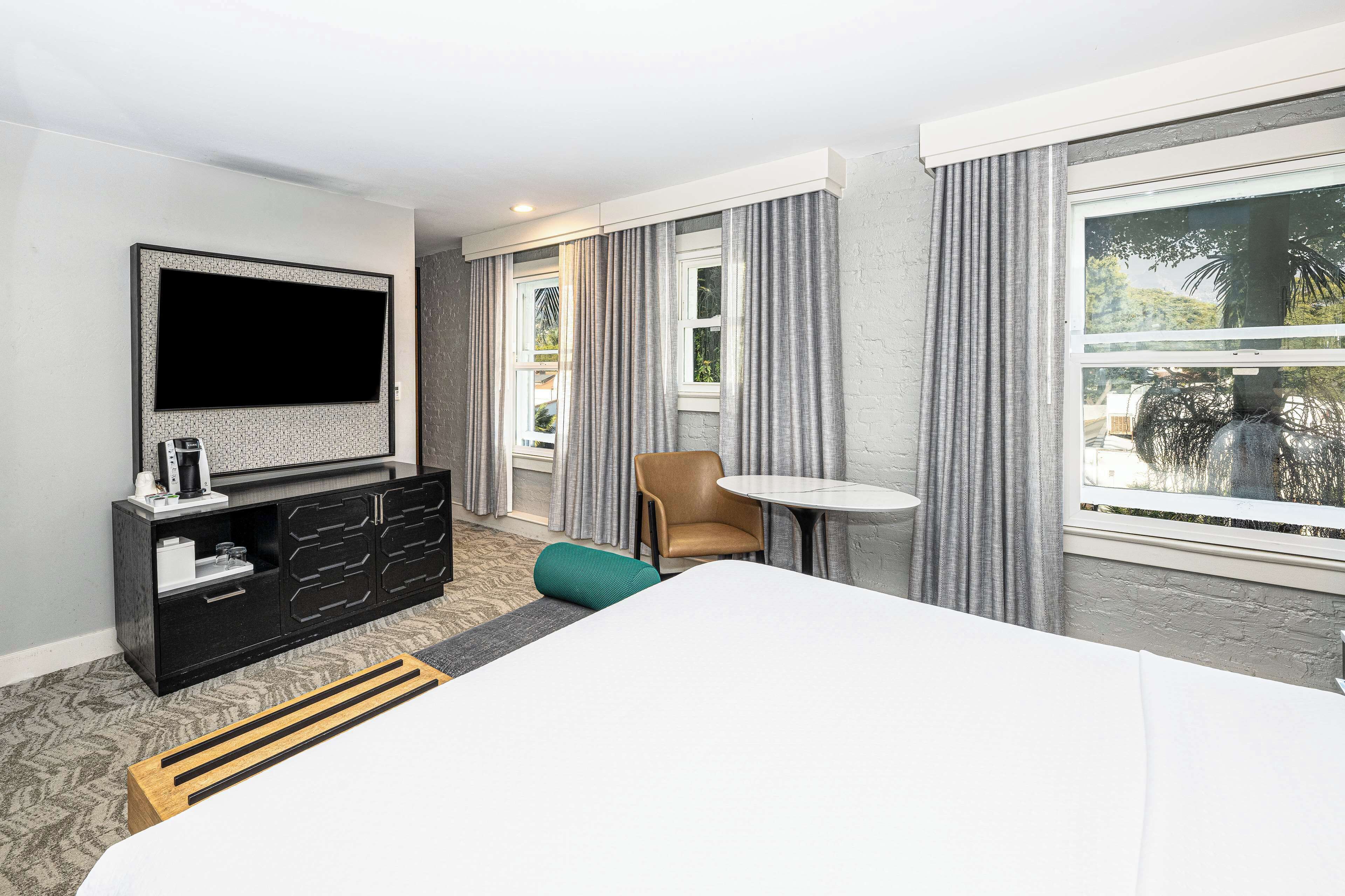 A hotel room with a bed, a TV, a small table, a chair, and a window with curtains.