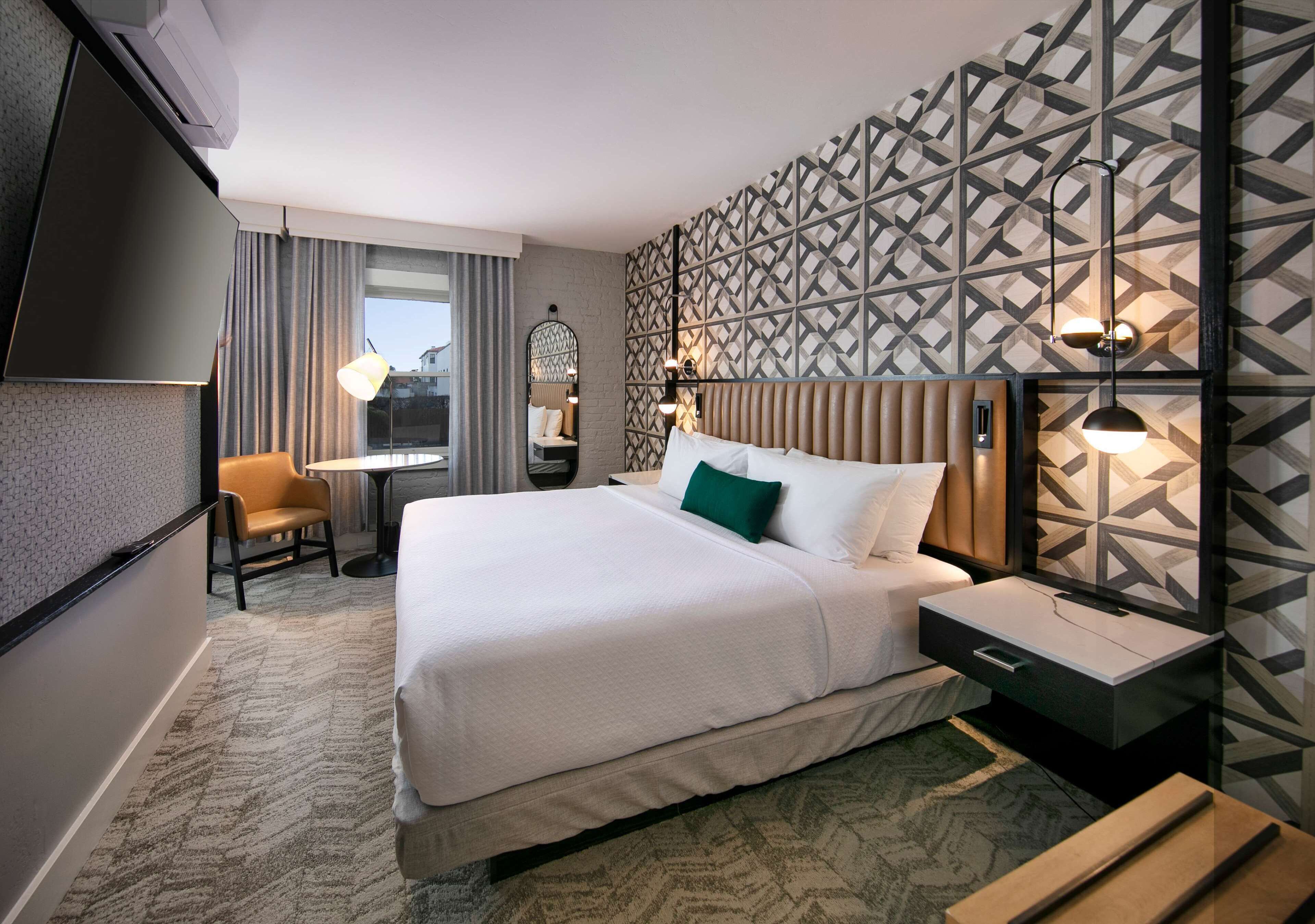 Ein Hotelzimmer mit einem großen Bett, einem Schreibtisch, einem Sessel und einem Fernseher, an der Wand ein Muster.