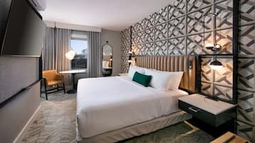 Ein Hotelzimmer mit einem großen Bett, einem Schreibtisch, einem Sessel und einem Fernseher, an der Wand ein Muster.