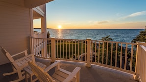 A balcony with white railings, a rocking chair, and a view of the ocean at sunset.