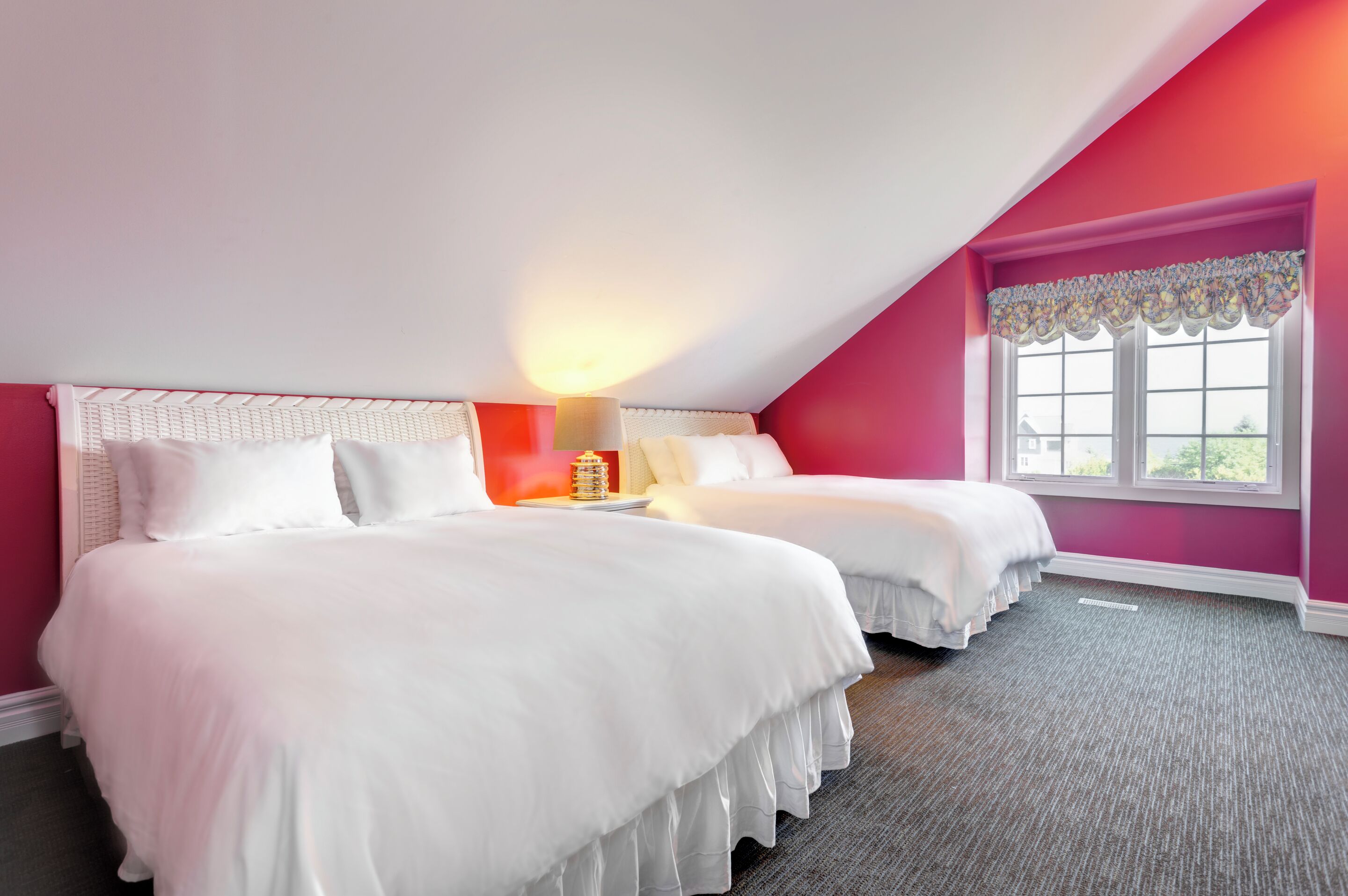 Un dormitorio con dos camas, una ventana con un adornado floral y una lámpara en la mesita de noche.