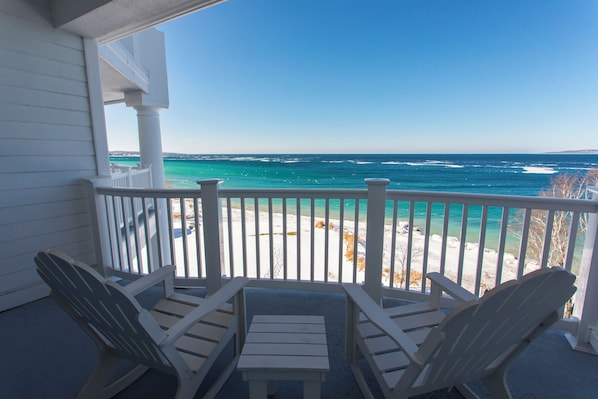 A balcony with white railings and two Adirondack chairs facing a beach with clear blue water.