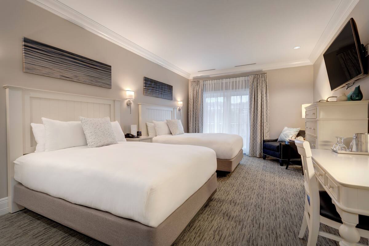 A hotel room with two beds, a desk, a chair, a TV, and a window with curtains.