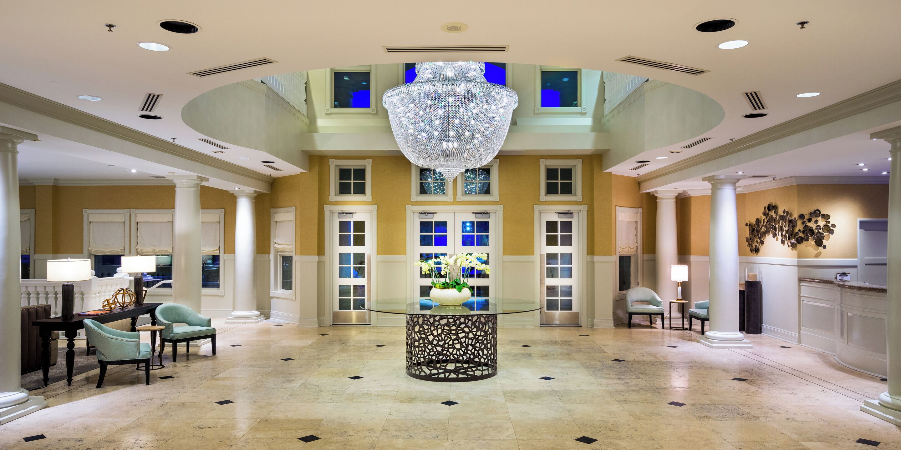 A spacious lobby with a central glass table, seating arrangements, and a decorative chandelier.