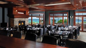 A restaurant with a fireplace, dining tables, chairs, and large windows with a view of the sea.