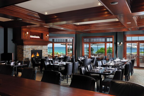 A restaurant with a fireplace, dining tables, chairs, and large windows with a view of the sea.