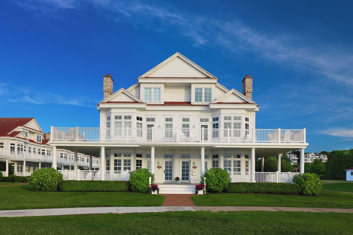 A large, two-story white building with a balcony, a covered front porch, and a brick chimney.