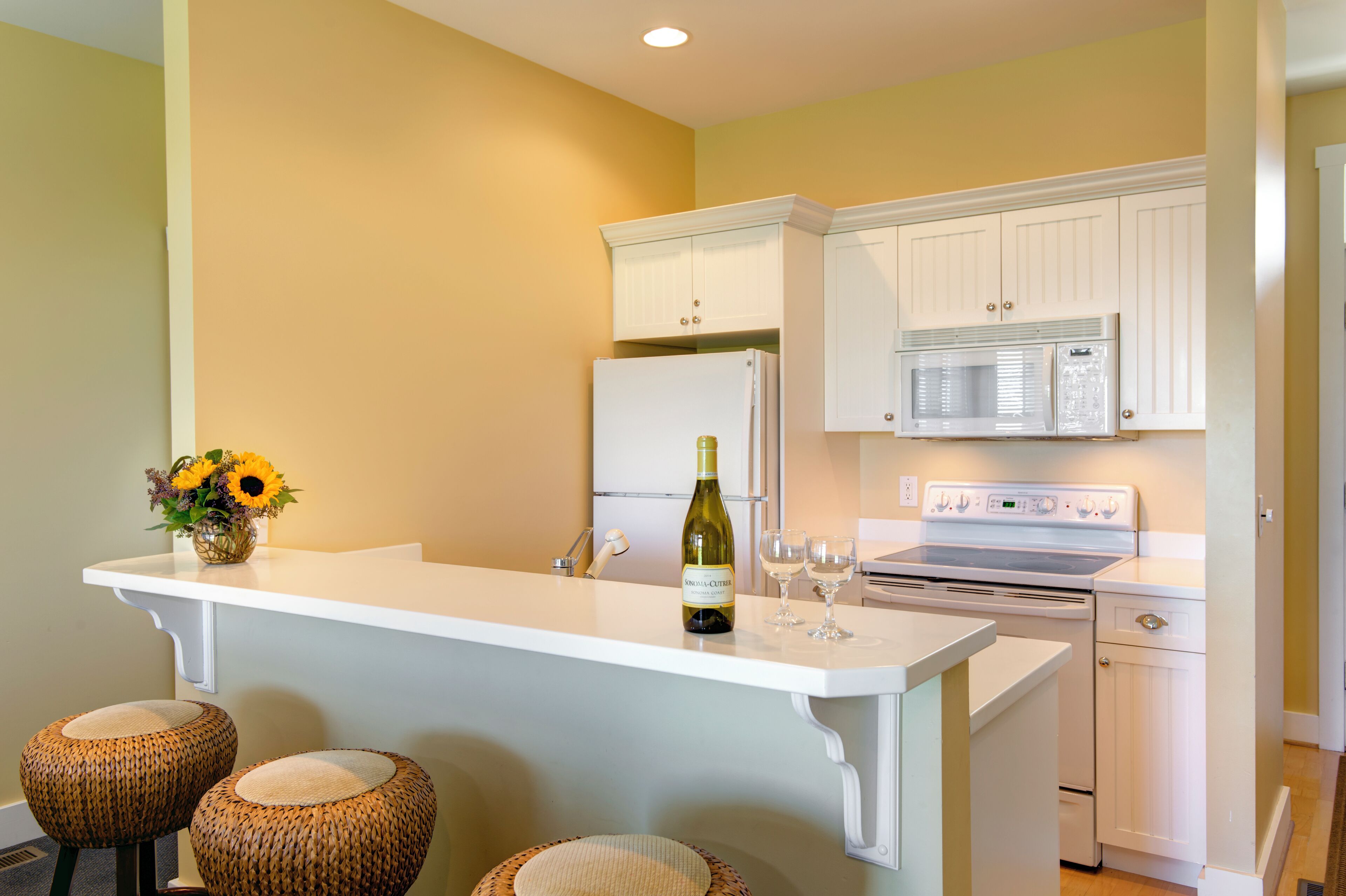 A kitchen with white cabinetry, a white countertop, a wine bottle, and two wicker stools.