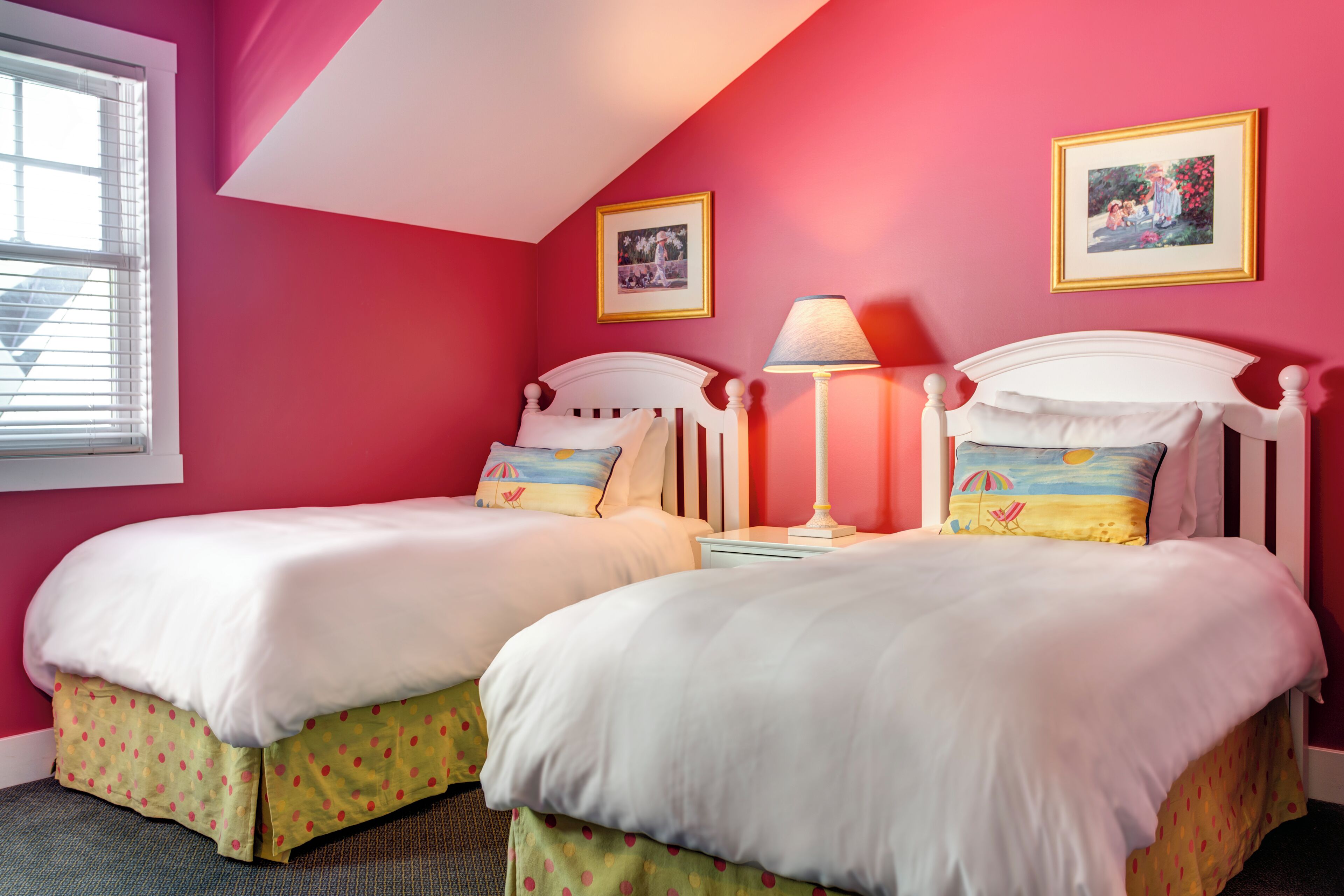 A room with two beds, each with a white headboard and matching bedding. There are two framed pictures on the walls, a window with blinds, and a bedside lamp on one of the beds.