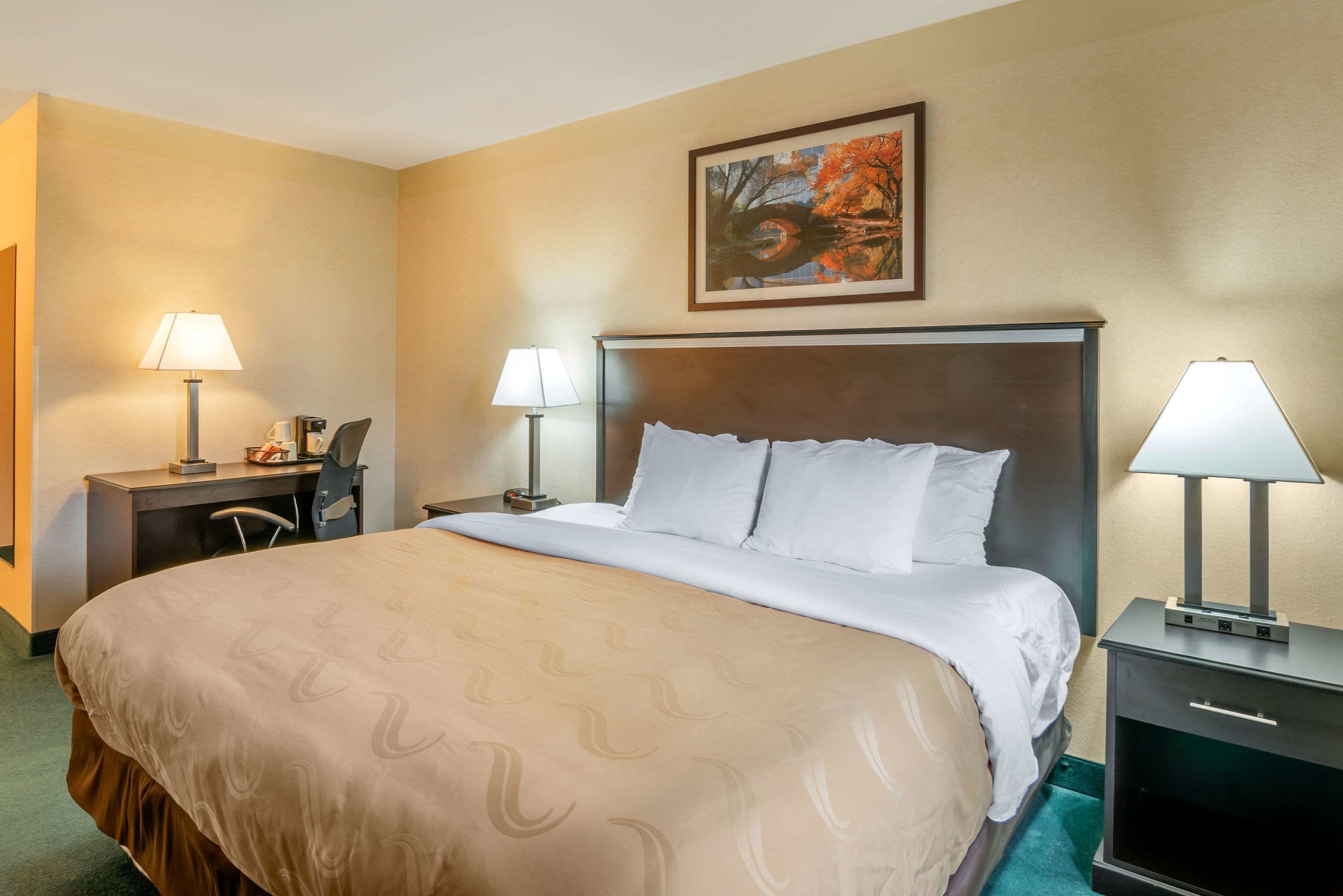 A hotel room with a large bed, two bedside tables with lamps, a desk with a chair, and a painting on the wall.