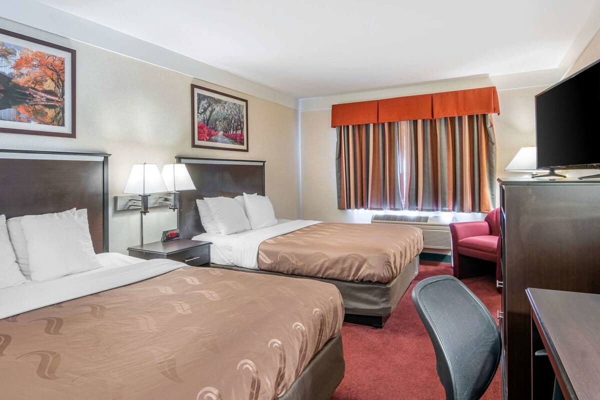 A hotel room with two beds, a desk, a chair, a TV, and a window with curtains.
