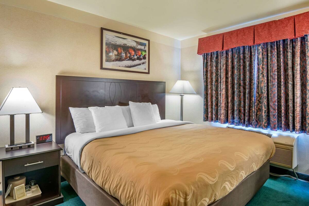 A hotel room with a large bed, two bedside lamps, a clock, a painting on the wall, and a window with patterned curtains.