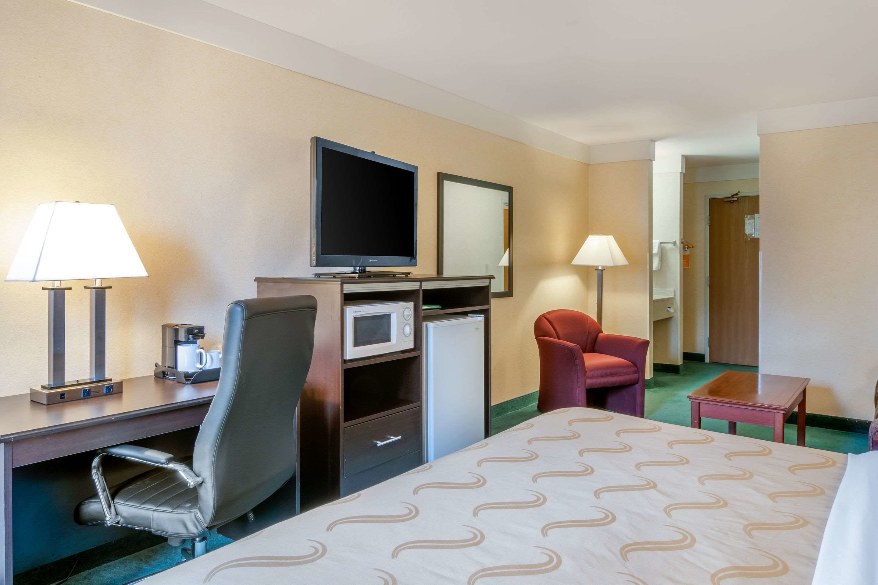 A hotel room with a bed, desk, chair, TV, microwave, and a small table.