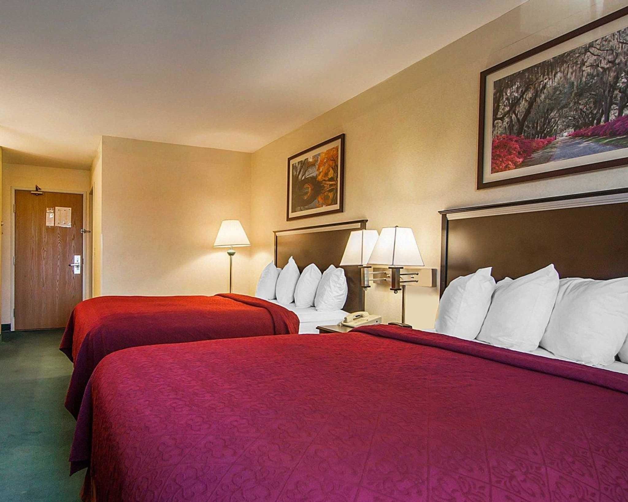 A hotel room with two beds, each with a red bedspread, white pillows, and bedside lamps. There is a painting on the wall above the beds and a door to the left.