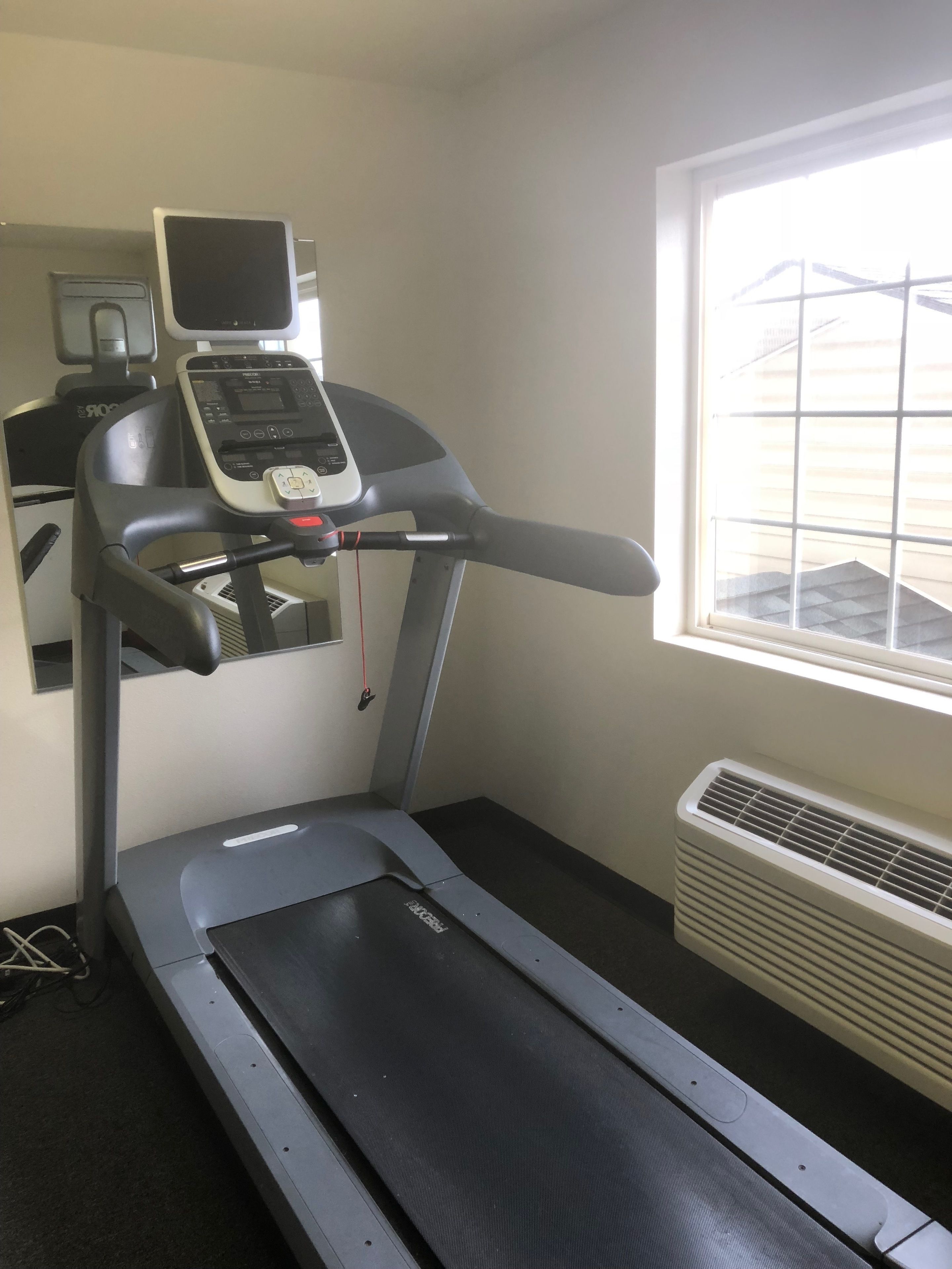 A treadmill in a room with a window, a wall-mounted TV, and a wall-mounted air conditioner.