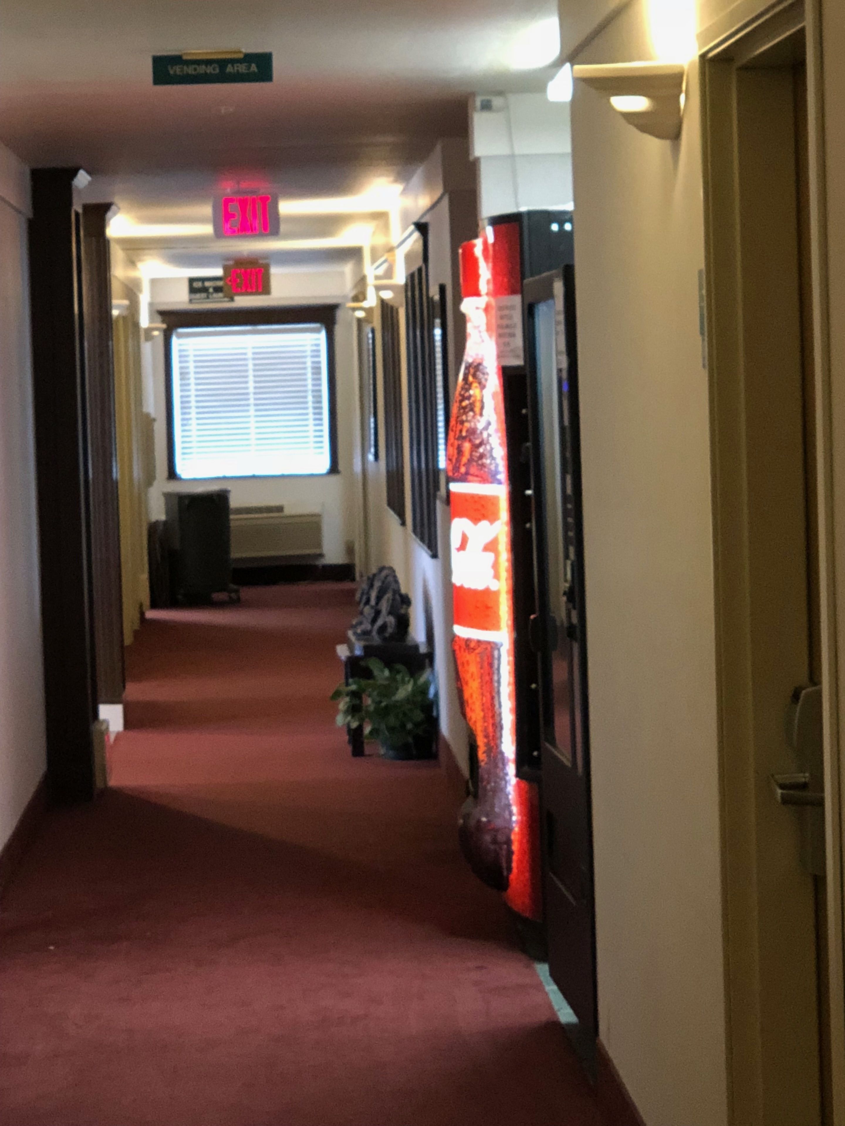 Corredor de hotel com tapete vermelho, área de máquinas de venda automática, placas de saída e uma máquina de Coca-Cola.