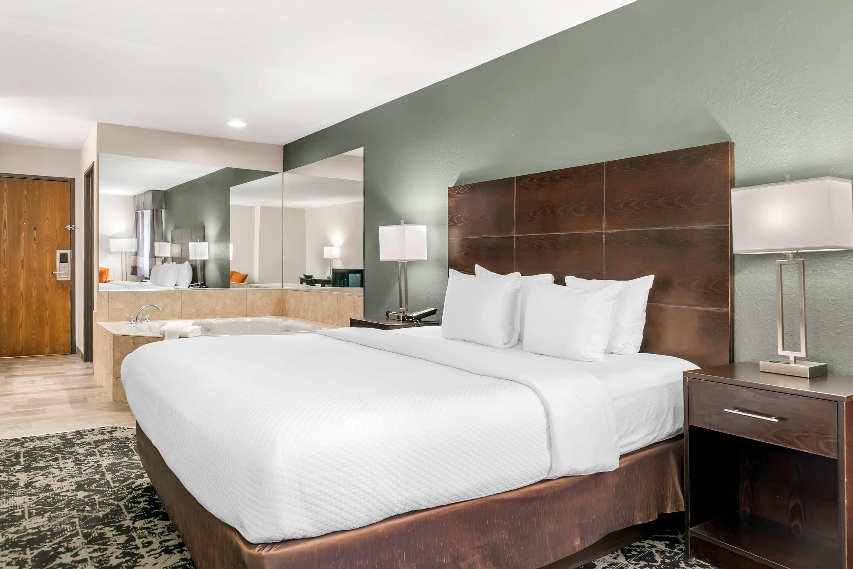 Habitación de hotel con una cama grande, una mesita de noche, un tocador, un espejo, un baño con lavamanos y bañera, y una puerta de madera.