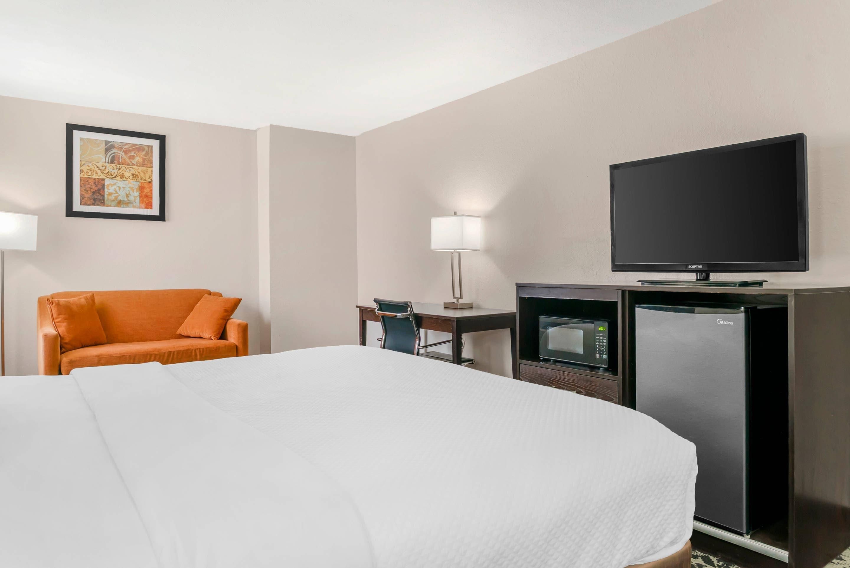 A hotel room with a bed, an orange chair, a desk with a lamp, a television, and a minibar.