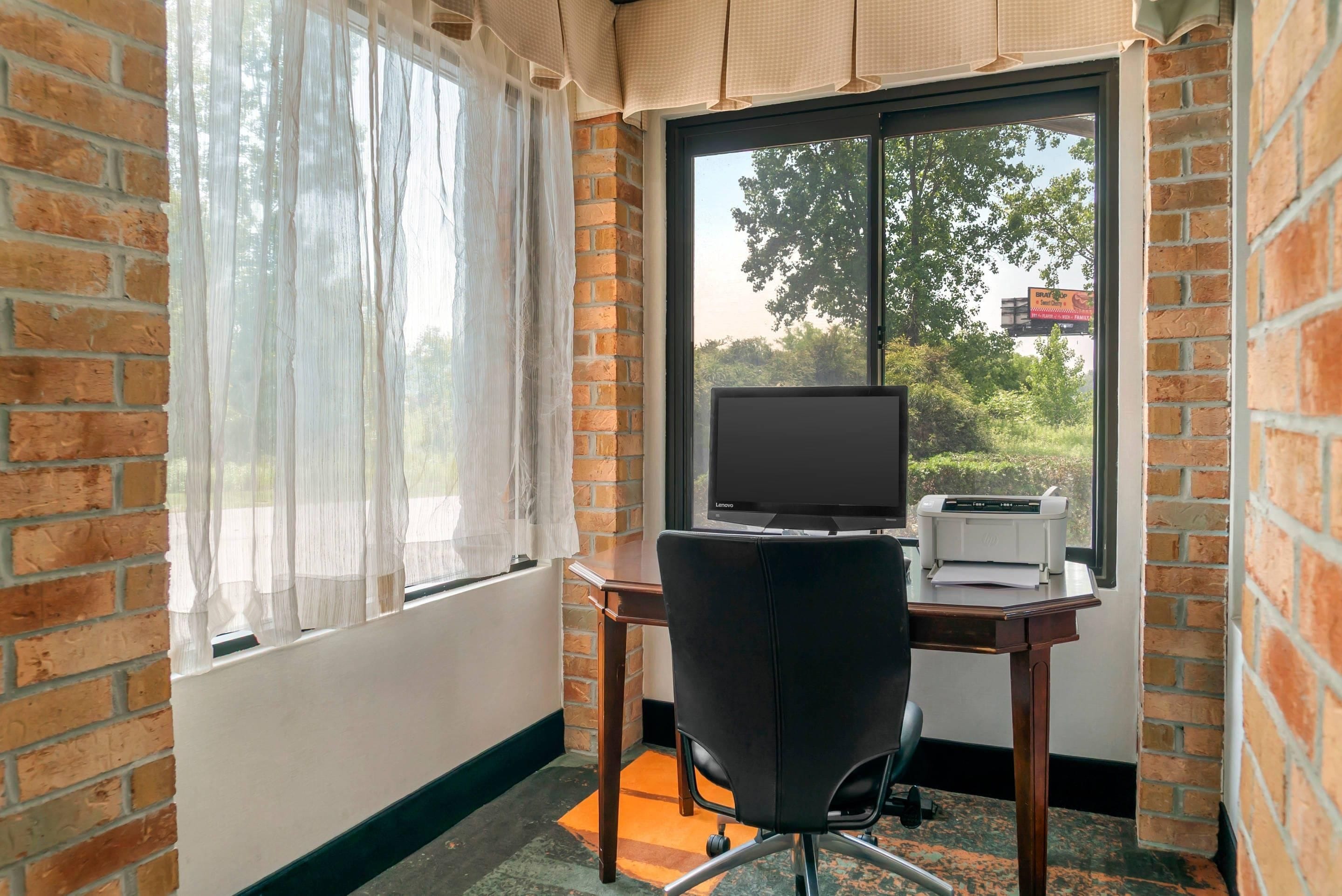 A room with a brick wall, a window with sheer curtains, a desk with a laptop and printer, and a black office chair.