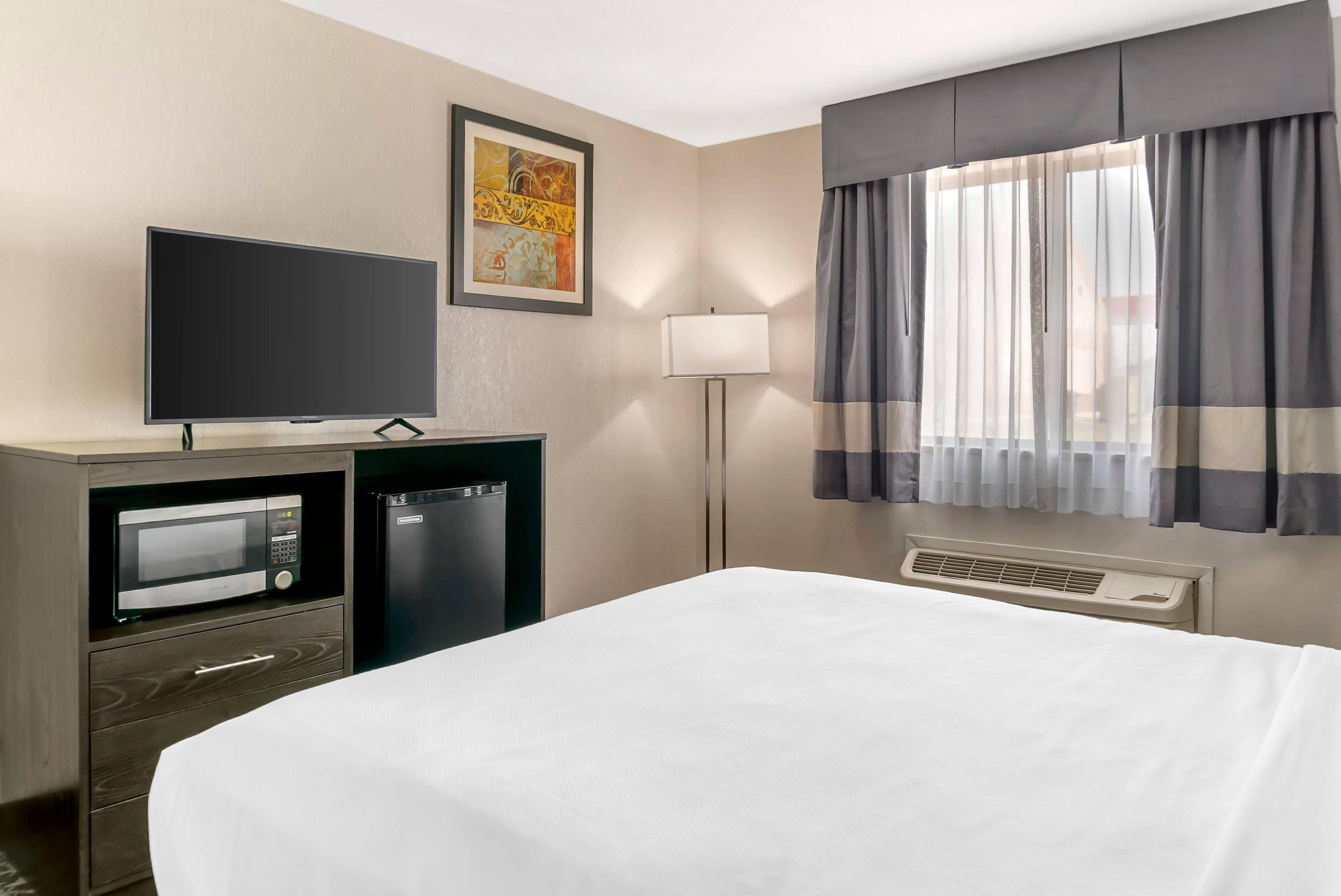 Ett hotellrum med en säng, en tv, en minibar, en lampa och ett fönster med gardiner.