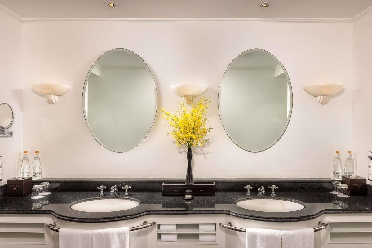 Une salle de bain moderne avec deux lavabos, un grand miroir et un vase rempli de fleurs jaunes.