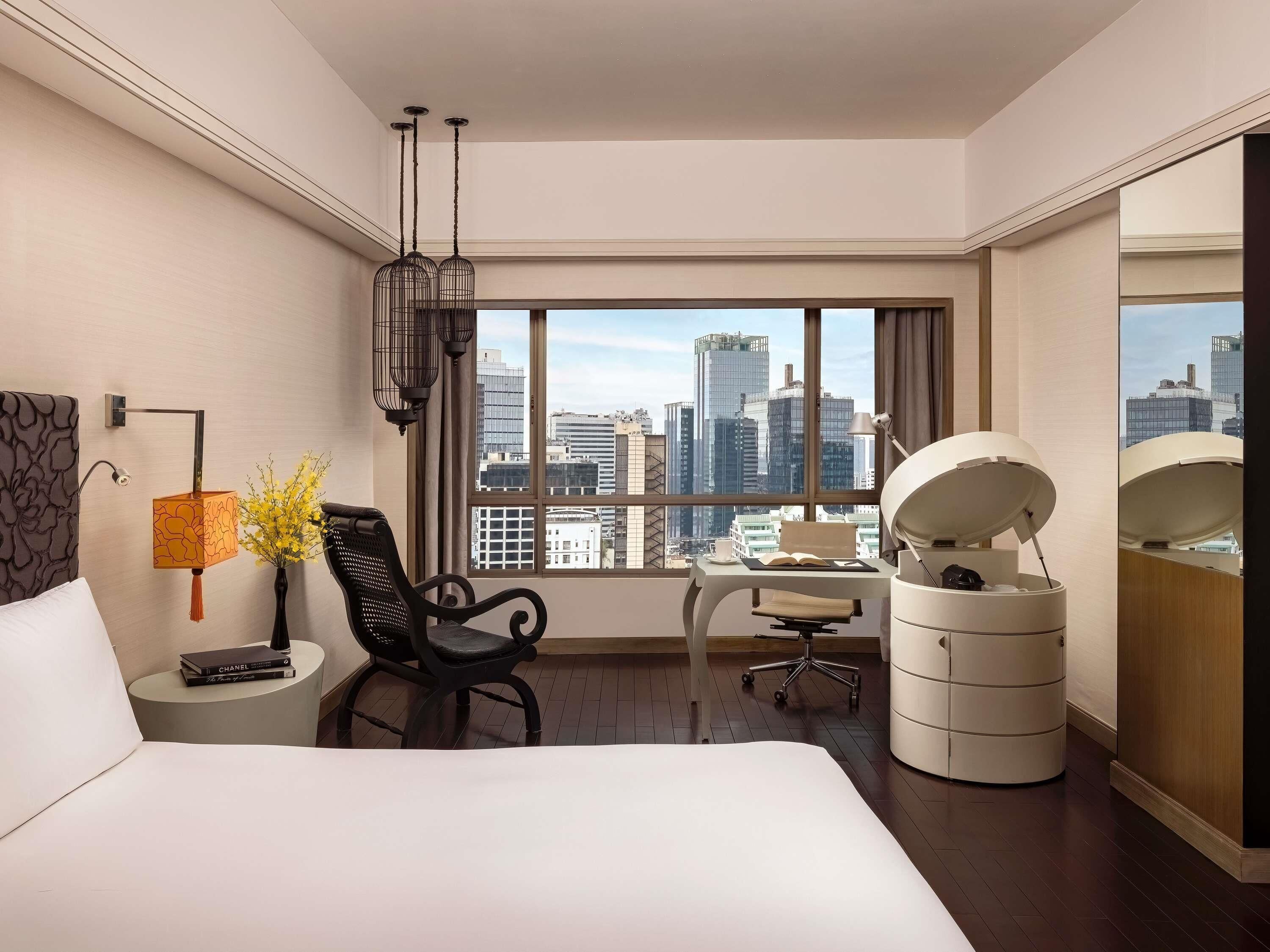 A modern hotel room with a large bed, a desk, a chair, and a view of the city skyline.