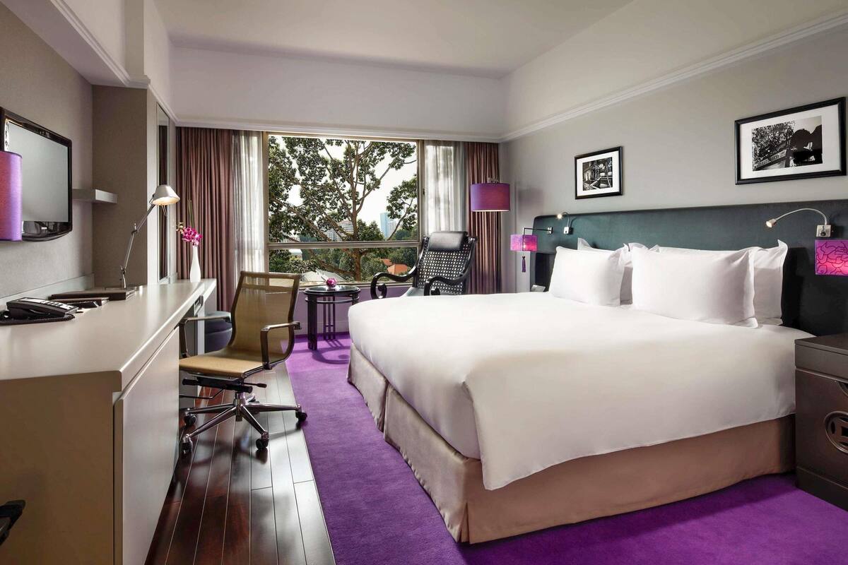 Une chambre d’hôtel avec un grand lit, un bureau, une chaise, une petite table et une fenêtre donnant sur des arbres et des immeubles.