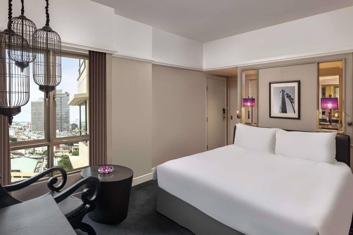 Une chambre d’hôtel avec un grand lit, un bureau, une chaise, une table ronde et une vue sur la ville grâce à de grandes fenêtres.