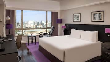 A hotel room with a large bed, a desk, a chair, and a view of the city.