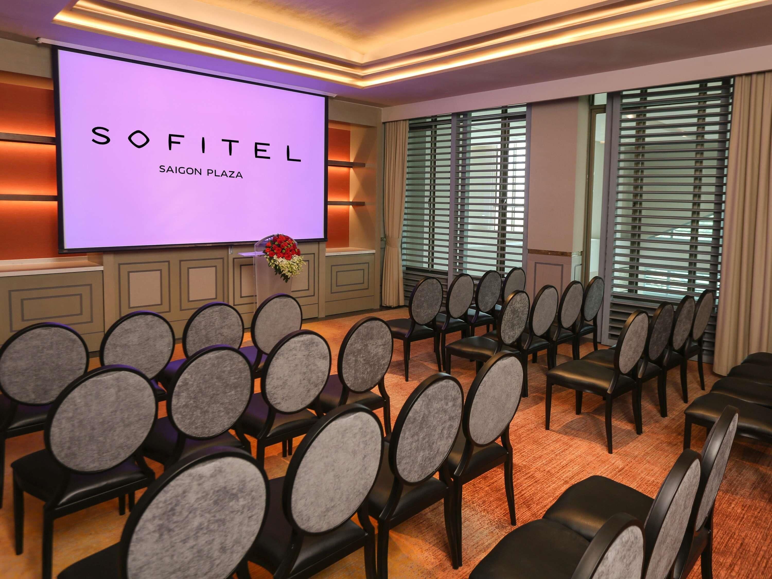 A conference room with a large screen displaying 'SOFITEL SAIGON PLAZA,' rows of chairs, and a window with blinds.