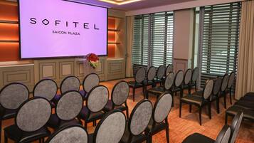A conference room with a large screen displaying 'SOFITEL SAIGON PLAZA,' rows of chairs, and a window with blinds.