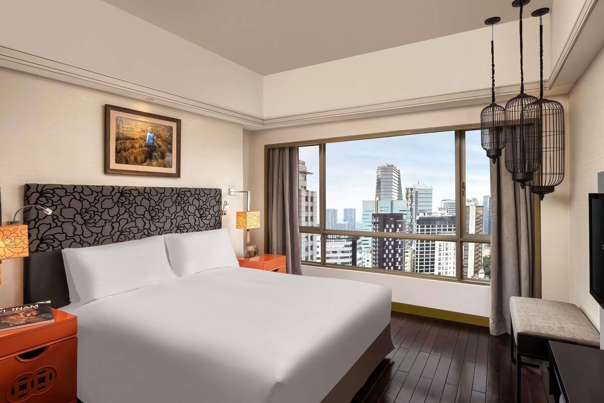 Une chambre d’hôtel avec un grand lit, une vue sur la ville, un tableau encadré et un lustre moderne.