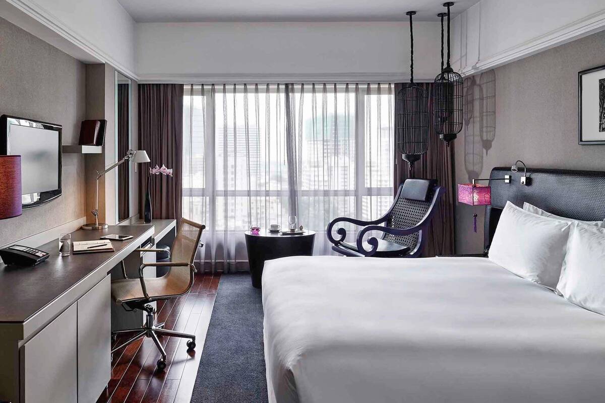 Una habitación de hotel moderna con una cama grande, un escritorio con silla, televisión y vistas de la ciudad a través de amplios ventanales.
