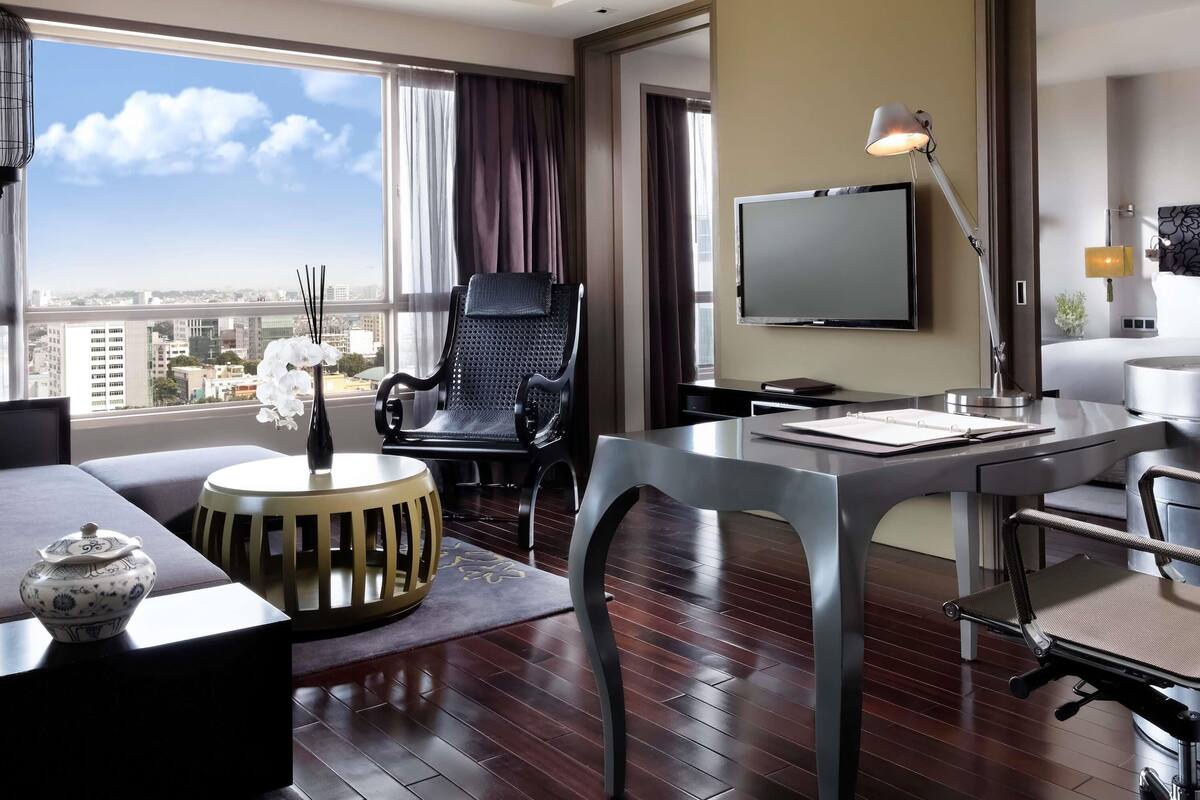 Una habitación de hotel moderna con un ventanal grande, un televisor de pantalla plana, un escritorio con una silla, un sofá y un jarrón decorativo.