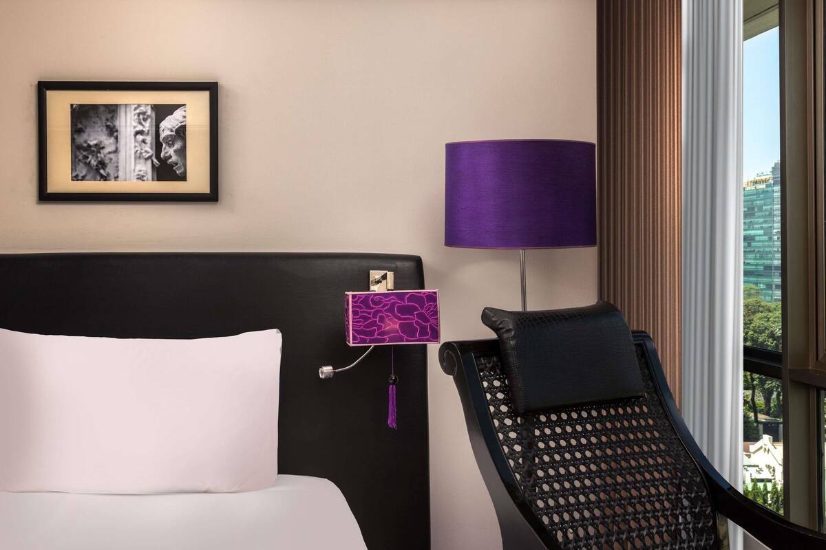 Habitación de hotel con cama, una silla, una lámpara, un cuadro enmarcado y una ventana con vistas.