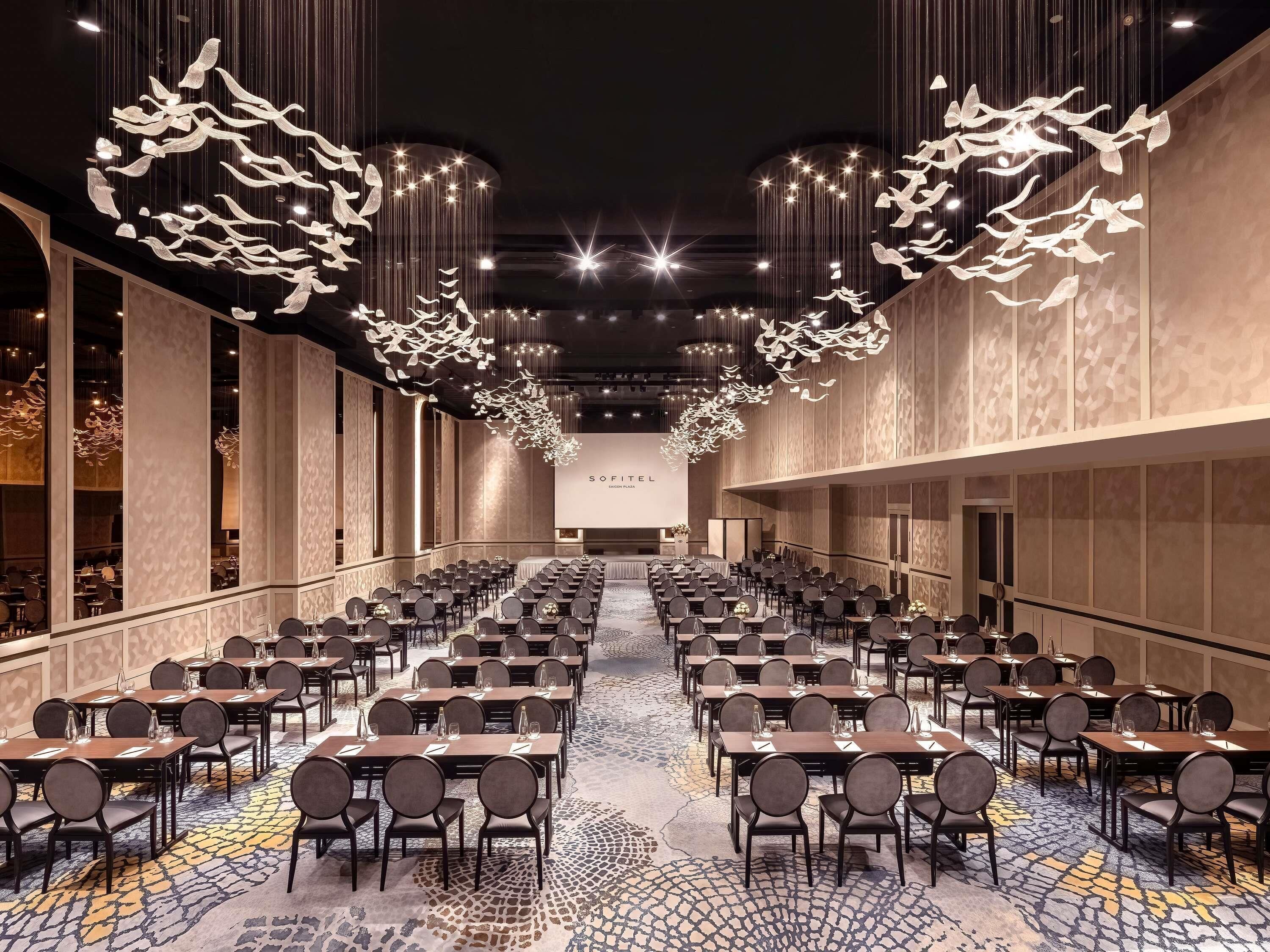 Une salle de conférence avec des rangées de tables et de chaises, une moquette à motifs et des luminaires suspendus.