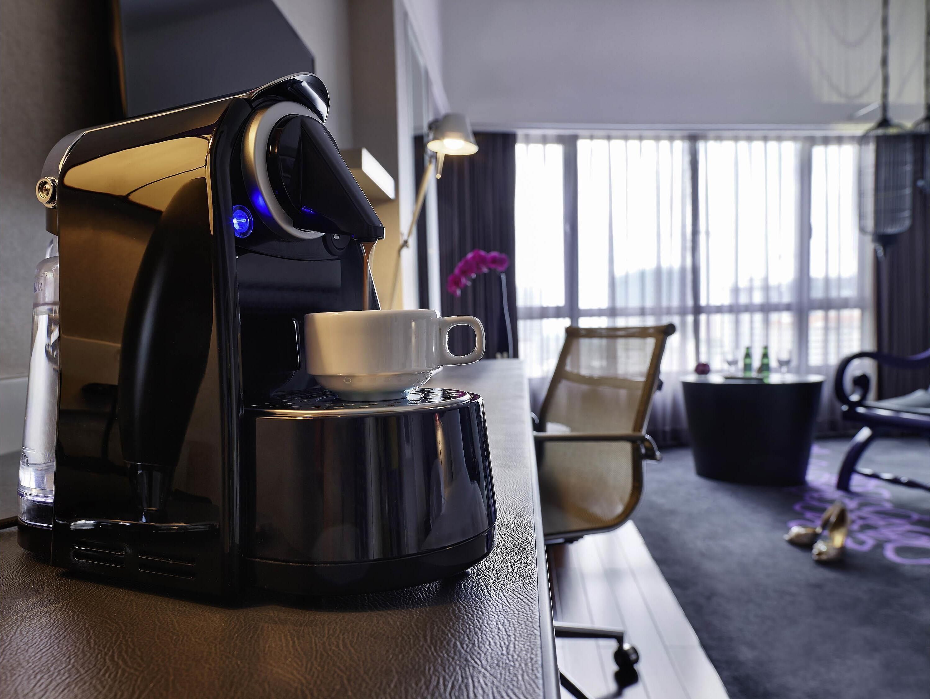 A coffee machine with a cup on a table, a chair, and a window with curtains.