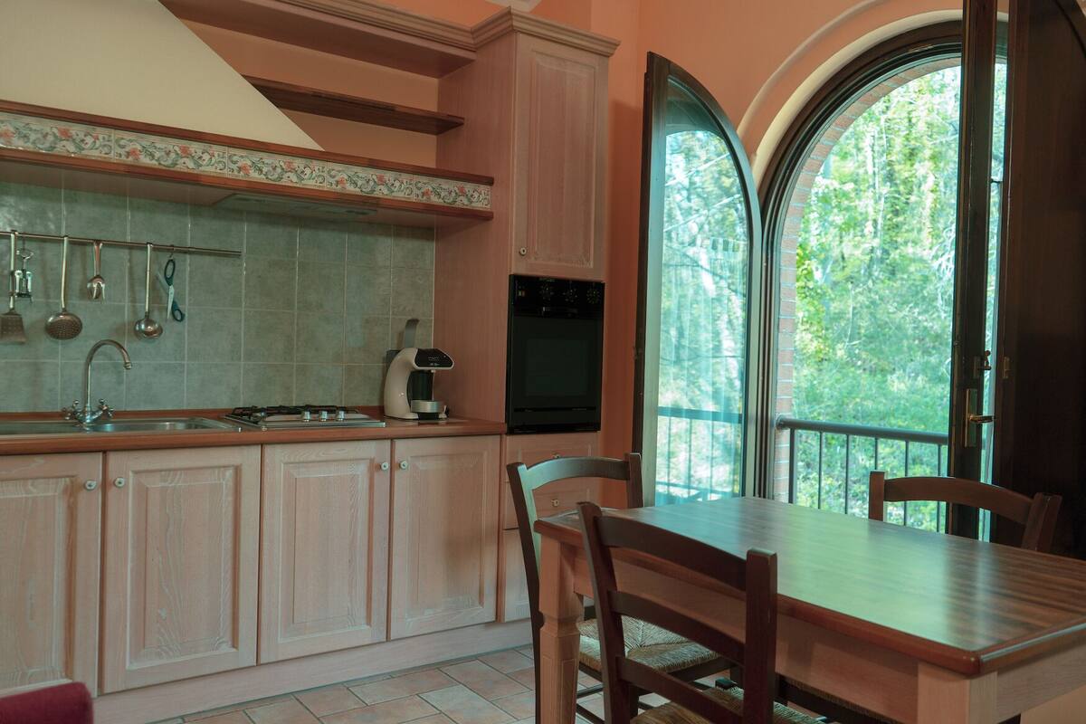 A kitchen with a dining area, a coffee maker, and a view of trees through a large window.